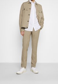 Beige Jacke mit zwei Brusttaschen und schwarzen Knöpfen, kombiniert mit einem weißen Hemd mit Punkten, beigen Hosen und weißen Sneakers. Minimalistisches Design.