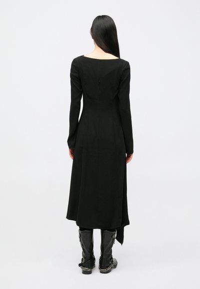 Μαύρο φόρεμα μακρυμάνικο με ίσια γραμμή, τετράγωνο ντεκολτέ, κλείσιμο με φερμουάρ στο πίσω μέρος και εφαρμοστή μέση που καταλήγει σε φούστα με ανοδικές πιέτες.