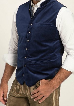 Mand iført en knappet marineblå fløjlsvest over en hvid opkrogede skjorte, med broderede brune traditionelle bukser.