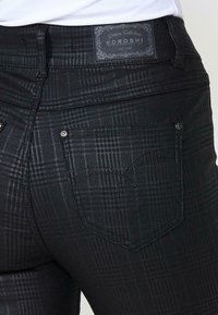 Jean à carreaux noirs avec poche arrière et étiquette en cuir portant la mention « Koroshi Denim Collection », porté avec un haut blanc.