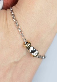Bracciale in catena d'argento con tre perline decorative: una dorata, un cuore argentato e una con pietre trasparenti. Texture liscia, design moderno.