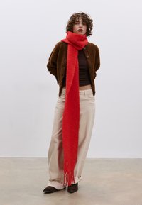 Červená nadýchaná šála přes tmavě hnědý kardigan. Světle béžové kalhoty a hnědé boty doplňují outfit. Rovná ofina na šále.