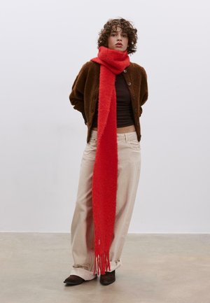 Sciarpa rossa e soffice drappeggiata su un cardigan marrone scuro. Pantaloni beige chiaro e scarpe marroni completano l'abbigliamento. Frangia dritta sulla sciarpa.