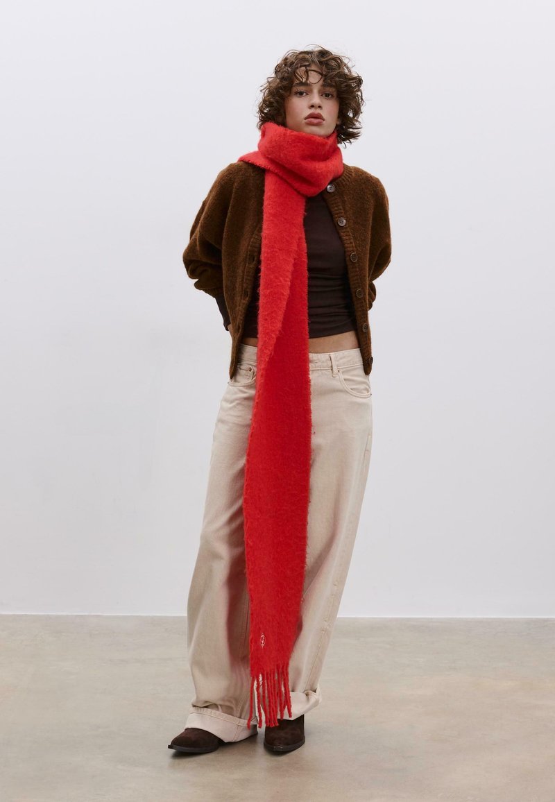 Červená nadýchaná šála přes tmavě hnědý kardigan. Světle béžové kalhoty a hnědé boty doplňují outfit. Rovná ofina na šále.