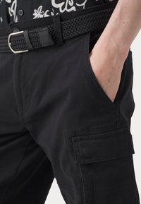 Personne portant un pantalon cargo noir avec une ceinture noire tressée et une chemise à fleurs, main placée dans la poche avant du pantalon.