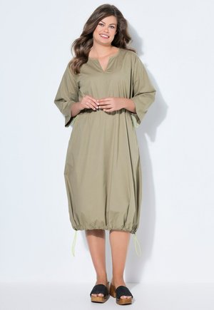 Femme aux longs cheveux bruns portant une robe ample olive verte jusqu'aux genoux avec un ourlet ajustable par cordon et des sandales à plateforme en bois noir, souriante.