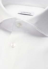 Chemise blanche à boutons avec un col pointu, montrant deux boutons et une étiquette de marque "seidensticker" visible à l'intérieur du col.