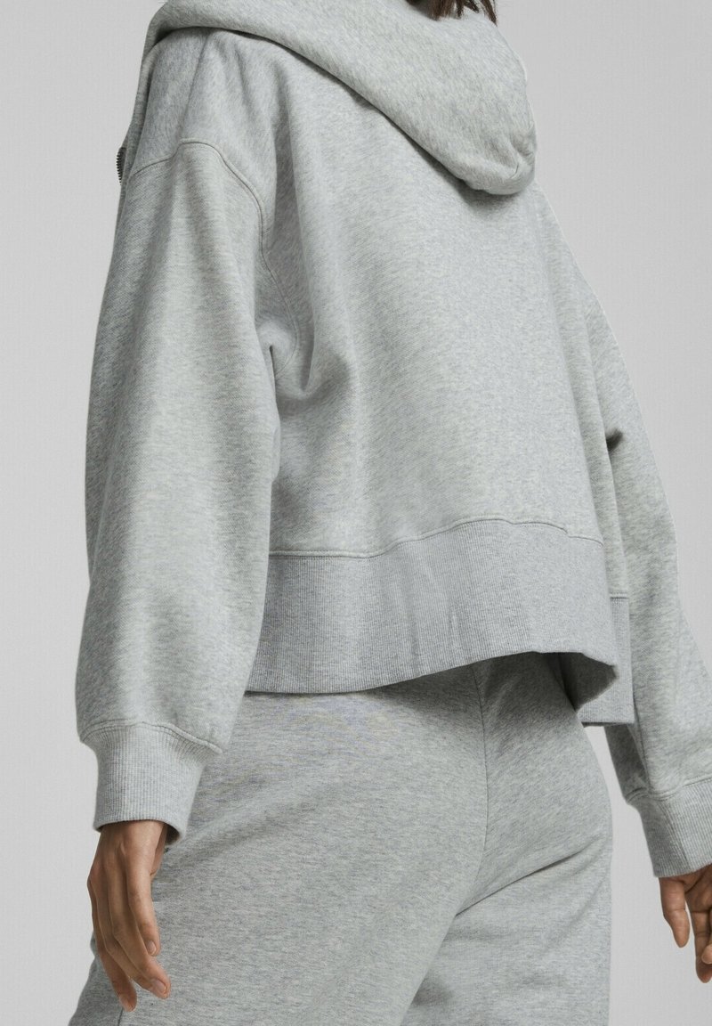 Personne portant un sweat à capuche ample gris clair et un pantalon de survêtement assorti, vue de dos de la taille vers le bas sur un fond neutre.