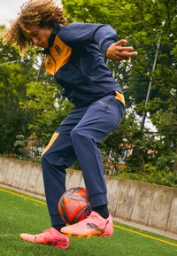 Blaue und orange Sportjacke sowie -hose; rosa Fußballschuhe; Spieler jongliert einen bunten Fußball auf grünem Rasen.