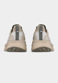 O pereche de pantofi sport bej cu tălpi groase, văzuți din spate, cu barete gri pentru prindere și un mic logo alb.