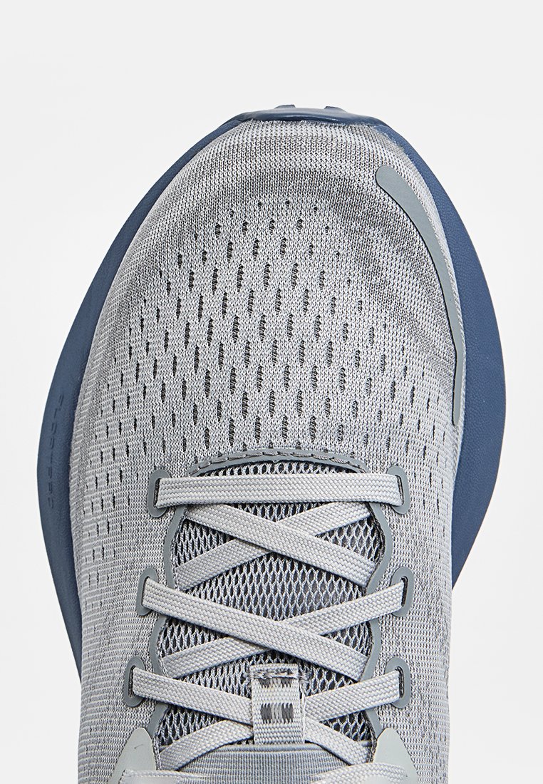 Zapatilla deportiva gris con parte superior de malla texturizada, punta redonda y cordones grises. La suela es azul oscuro con un acabado liso.