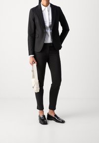 Blazer noir avec revers, chemise blanche à col, pantalon noir slim et mocassins noirs brillants. Porte un sac à bandoulière blanc.