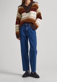 Suéter de punto con cuello, con franjas marrones, naranjas, blancas y rojas. Combinado con jeans de mezclilla azul de cintura alta y zapatos negros.