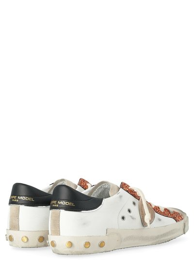 Sneakers bianchi a basso profilo con dettagli neri sul tallone, accenti di glitter arancioni, lacci beige e borchie in ottone sui bordi della suola, con segni di usura.