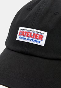 Černá baseballová čepice s přišitou bílou nášivkou s modrým a červeným textem, na které je napsáno "LATELIER Garage Workshop", vyrobená z hladké, texturované látky.