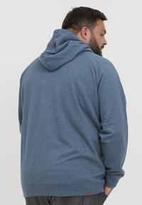 Sudadera con capucha en azul, hecha de tela suave. Presenta puños y dobladillo de canalé, con un detalle de etiqueta de cuero en la parte inferior. Diseño simple y casual.