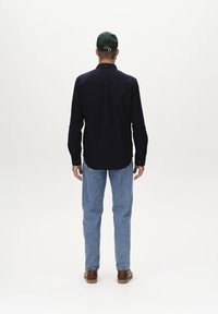 Camicia blu navy con maniche lunghe, abbinata a jeans azzurri e scarpe marroni, vista da dietro su uno sfondo neutro.