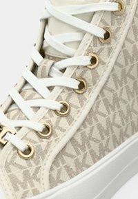 Κοντινό πλάνο μπεζ ψηλού παπουτσιού με λευκά κορδόνια, χρυσά τιράντες και ύφασμα με μοτίβο που επαναλαμβάνει τα γράμματα "MK".