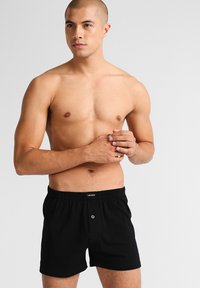Boxers de algodón negros con una cinturilla elástica y detalle de botón en la parte frontal, llevados por un modelo con la cabeza rapada y torso desnudo.