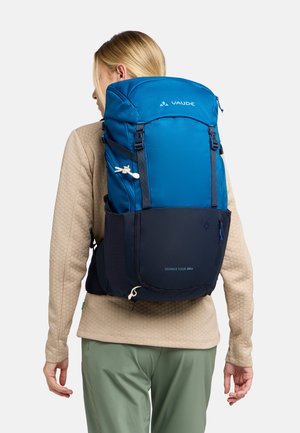 Zaino Vaude Skomer Tour 34+ blu, con un cappuccio superiore, spallacci regolabili e un pannello inferiore blu scuro dotato di anelli per accessori e una finitura testurizzata.