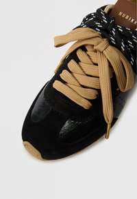 Zapatilla negra con texturas de ante y cuero brillante, con cordones beige y acentos dorados en los ojetes. Punta redondeada y cuello acolchado.