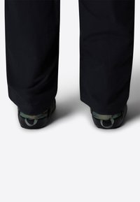 Pantaloni neri abbinati a scarpe mimetiche verde scuro, con superficie testurizzata e cinturini regolabili per il tallone.