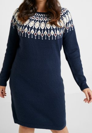 Woman wearing navy blue knit sweater dress with white, beige, and brown geometric pattern across chest and shoulders.