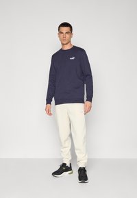 Sweatshirt à manches longues navy avec un logo blanc, associé à un pantalon de survêtement crème et des baskets noires avec des accents verts. Design simple avec des poignets côtelés.