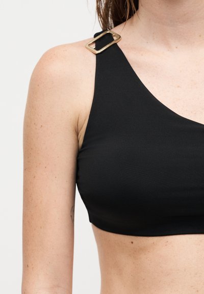 Μαύρο ασύμμετρο πάνω μέρος μπικίνι με χρυσή αγκράφα στον μονό ιμάντα του ώμου, που φορά ένα άτομο με ανοιχτόχρωμο δέρμα, σε απλό φόντο.