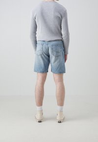 Persona vestida con una camiseta de manga larga de canalé gris claro, pantalones cortos de mezclilla azul claro, calcetines blancos y zapatillas de caña alta beige, de pie de espaldas.