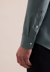 Langarmshirt in een gedempte groene stof. Heeft knoopmanchetten met witte knopen en een klein geborduurd ontwerp op de mouw.