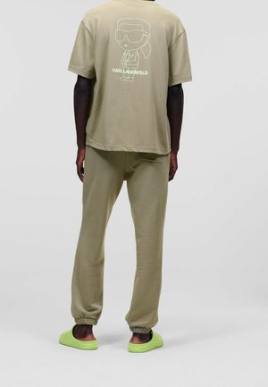 Khaki T-Shirt mit Cartoon-Grafik auf dem Rücken, kombiniert mit passenden Jogginghosen mit elastischen Bündchen. Die Schuhe sind grüne Slipper.
