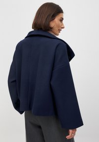 Tumedast sinine lühike jakk suure kraega, laiad varrukad ja tekstureeritud kangas. Omab lihtsat, minimalisti disaini ilma kaunistusteta.