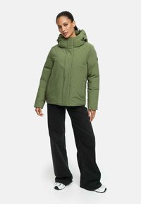 Olijfgroene pufferjack met een hoge kraag en een verstelbare capuchon. Glad textuur, ritssluiting aan de voorkant, en zijzakken. Gecombineerd met zwarte wijde broek.