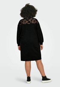 Vestido negro con un respaldo acentuado de encaje, mangas largas y un corte relajado, combinado con zapatillas negras altas. Tela texturada y diseño sencillo.