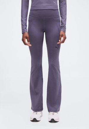 Personne portant des leggings évasés violets ajustés avec poches latérales et des chaussures de sport blanches sur un fond uni.