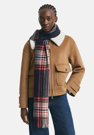 Giacca di lana marrone con colletto in finta pelliccia bianca, dotata di due tasche a pattina. Sciarpa a quadri blu navy con accenti rossi drappeggiata attorno al collo.