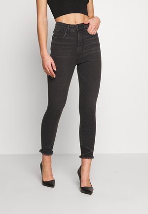 Femme portant un jean skinny noir taille haute avec des ourlets effilochés et des escarpins noirs à bouts pointus, debout devant un fond blanc uni.