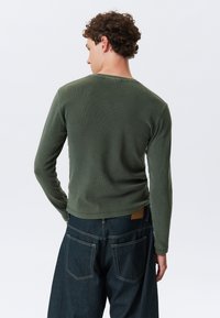 Langermet, olivengrønn skjorte laget av ribbestrikket stoff, med rund hals og rett kant, i kombinasjon med mørke denimjeans.