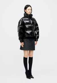 Γυναίκα ντυμένη με μια γυαλιστερή μαύρη μπουφάν, ανθρακί μίνι φούστα, μαύρες κάλτσες μέχρι το γόνατο και μαύρες γόβες με μυτερή μύτη, να στέκεται μπροστά σε λευκό φόντο.