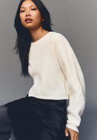 Bijeli teksturirani džemper opuštenog kroja s spuštenim ramenima, u kombinaciji s lepršavom crnom suknjom na neutralnoj pozadini.