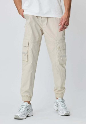 Homme portant un pantalon cargo beige avec des bords élastiques et des baskets blanches New Balance sur un fond clair uni.