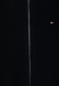 Maglione a maglia nera con zip, realizzato in tessuto testurizzato. Presenta una zip argento e un piccolo logo con dettagli in rosso, bianco e blu.