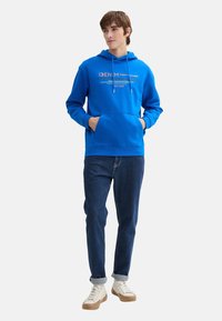 Felpa blu in cotone con stampa del logo, cappuccio regolabile con coulisse, tasca a marsupio, abbinata a jeans scuri e scarpe da ginnastica chiare.