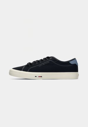 Zwarte canvas sneaker met witte zool, blauwe accenten op de hak, ronde neus, zwarte veters en contrasterende stiksels. Bevat het merkelogo aan de zijkant.