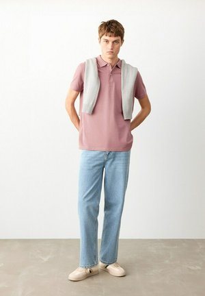 Młody mężczyzna stoi z rękami za plecami, ubrany w różową koszulkę polo, jasnoszary sweter zarzucony na ramiona, jasnoniebieskie dżinsy i beżowe trampki.