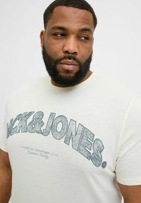 Kreemikas puuvillane T-särk sinise kulunud "JACK & JONES" graafikaga ees. Lühikesed varrukad ja ümmargune kaelus. Pehme tekstuur.