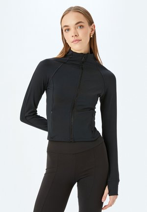 Svart zip-up jacka med långa ärmar, tillverkad av stretchigt tyg. Har en nära passform och tumhål vid ärmsluten. Slät textur, kort design.