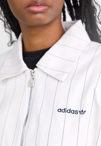 Balta juostelėmis dryžuota striukė, pagaminta iš lygaus audinio, su užtrauktuku ir navy adidas logotipu kairiajame šone.