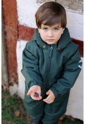 Lapsi vihreässä vedenpitävässä haalariasussa pitää avoimessa kämmenessään kahta kastematoa esitellen asun materiaalia ja design-ominaisuuksia.